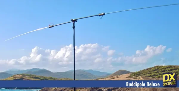A Buddipole Deluxe portable dipole on a telescoping mast, set up outdoors against a blue sky with hills and coastline behind it.